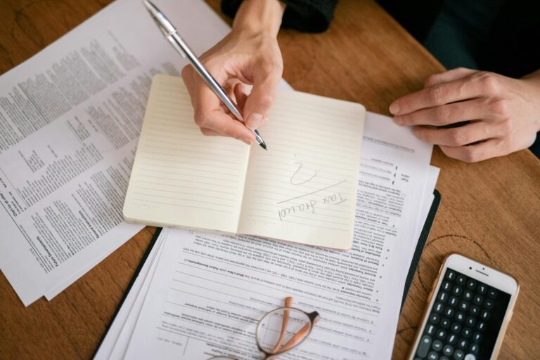 Close-up of hands writing on a notebook surrounded by tax documents and a smartphone.